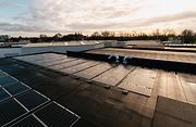 Zonnepanelen op een plat dak onder een bewolkte hemel bij zonsondergang, met bomen en gebouwen op de achtergrond.